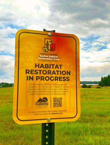 A yellow Alabama State Parks sign in a grassy field reads “Habitat Restoration in Progress,” with a Native Habitat Restoration Crew logo, QR code, and information about protecting native habitats.