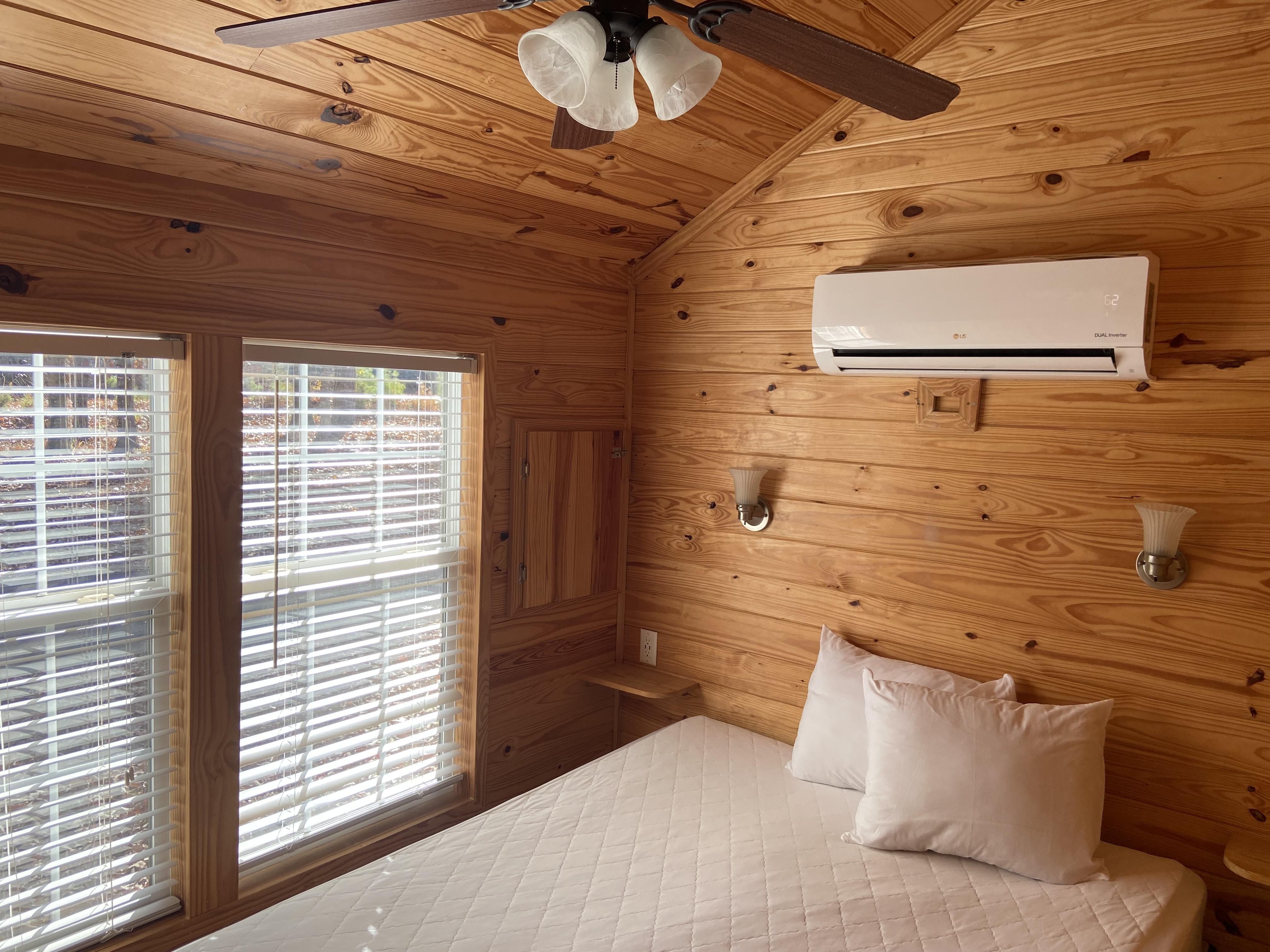 Queen bed with ceiling fan in a room with wooden walls and two windows