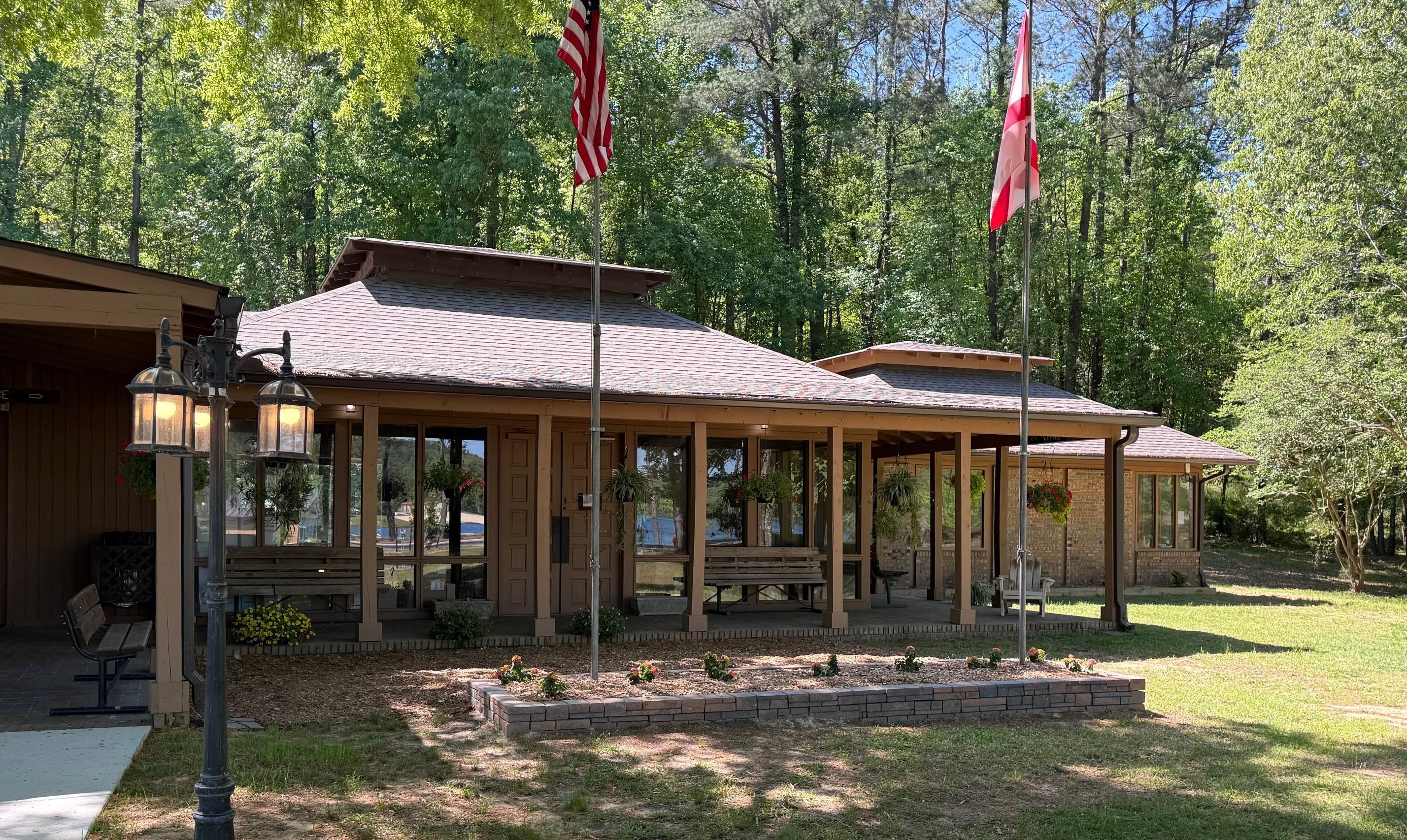 A single-story lodge-style building at Lake Lurleen State Park with a wide covered porch sits in a wooded area. Two flagpoles in front display the American flag and the Alabama state flag. Hanging plants and benches line the porch, while a small landscaped flower bed and a lamppost are in the foreground. Sunlight filters through tall trees surrounding the building.