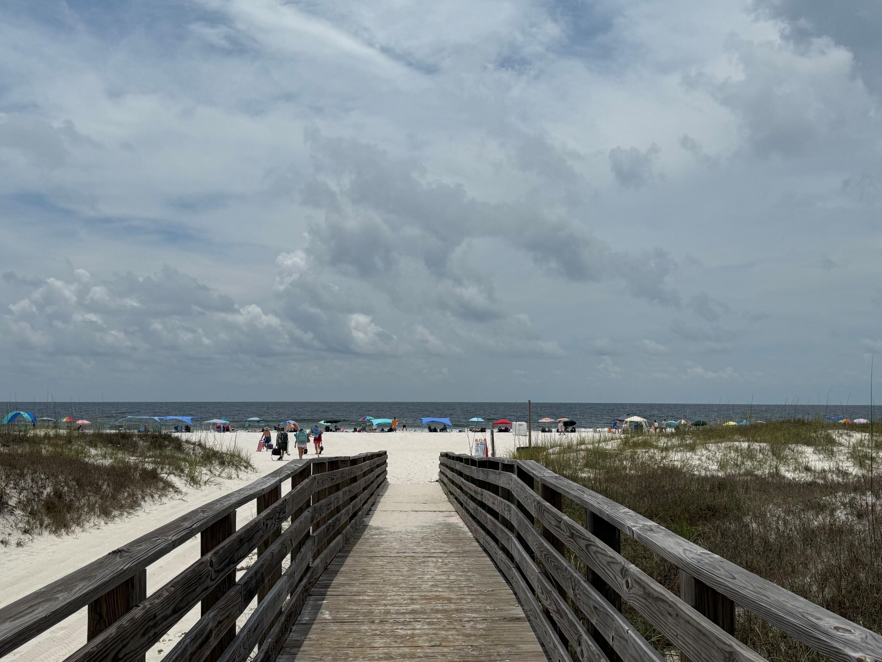 A wooden boardwalk leads over sand dunes to a wide beach lined with colorful umbrellas and beachgoers. The sky is mostly cloudy, and the ocean is visible in the distance.