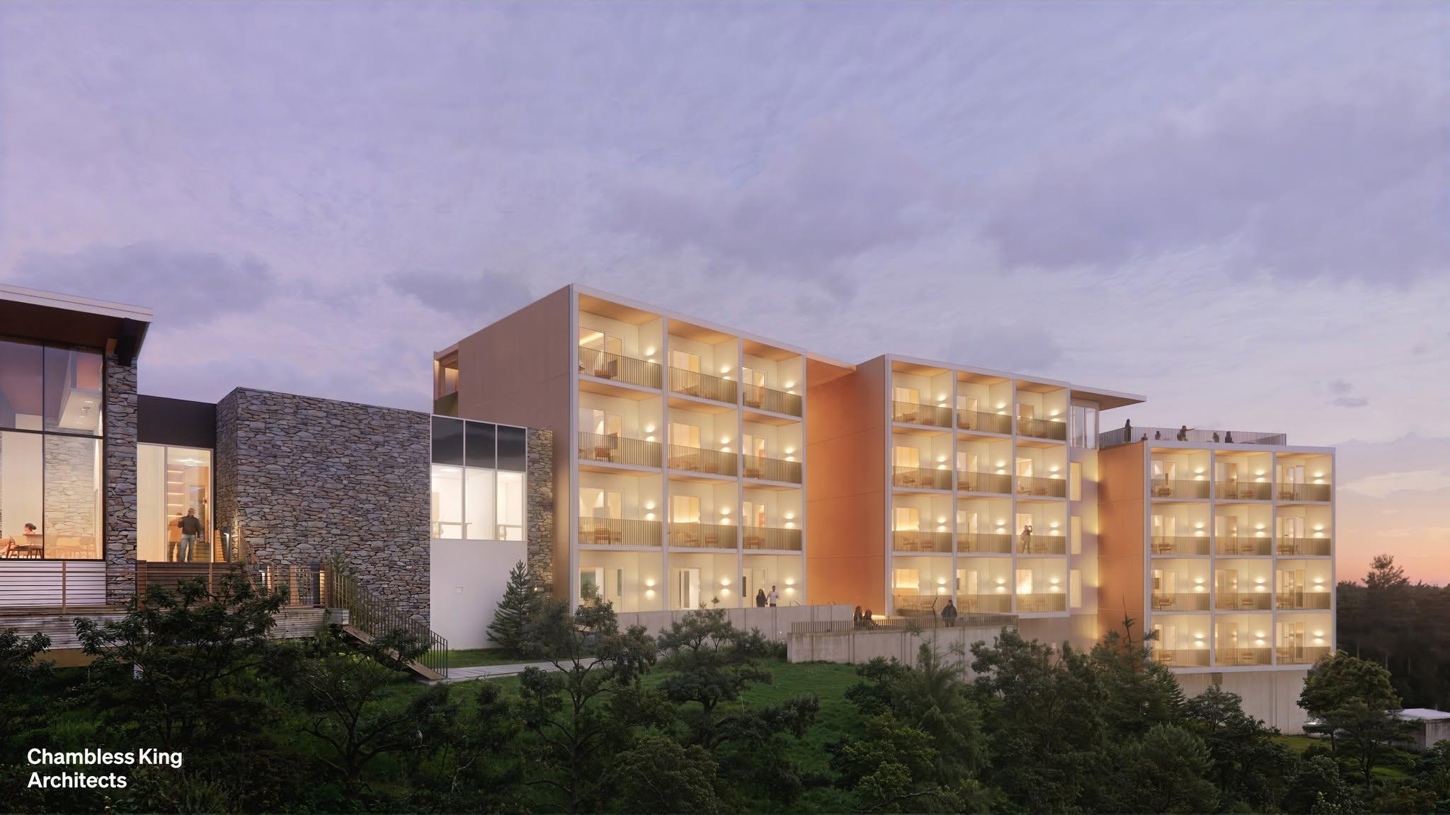  A modern multi‑story building with a warm, glowing interior stands on a hillside at dusk. The structure features several stacked rectangular sections, each with rows of illuminated balconies facing outward. To the left, an attached building is clad in stone with large windows. Trees and greenery fill the lower portion of the scene, and the sky above is soft purple with scattered clouds. The bottom-left corner includes small white text that reads “Chambless King Architects.”