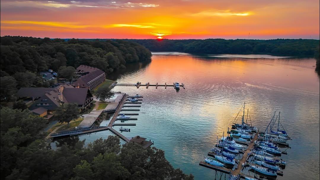 A vibrant sunset casts orange, pink, and purple colors across a calm lake surrounded by dense green trees. A marina with numerous docked sailboats sits on the right side, while lodge-style buildings and smaller docks line the left shoreline. The sun is just above the horizon, reflecting brightly on the water, creating a serene and picturesque scene.