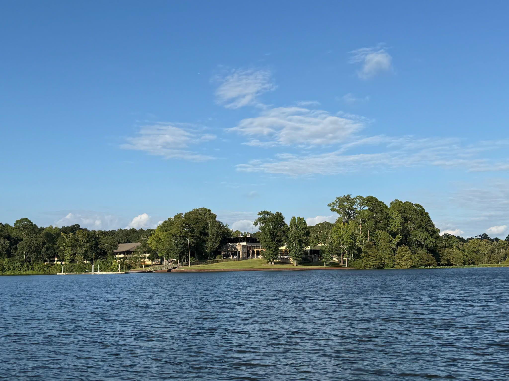 A calm lake stretches across the foreground under a clear blue sky with scattered white clouds. Along the distant shoreline, several buildings sit partially hidden among dense green trees. The water is gently rippled, and the overall scene appears peaceful and sunlit.