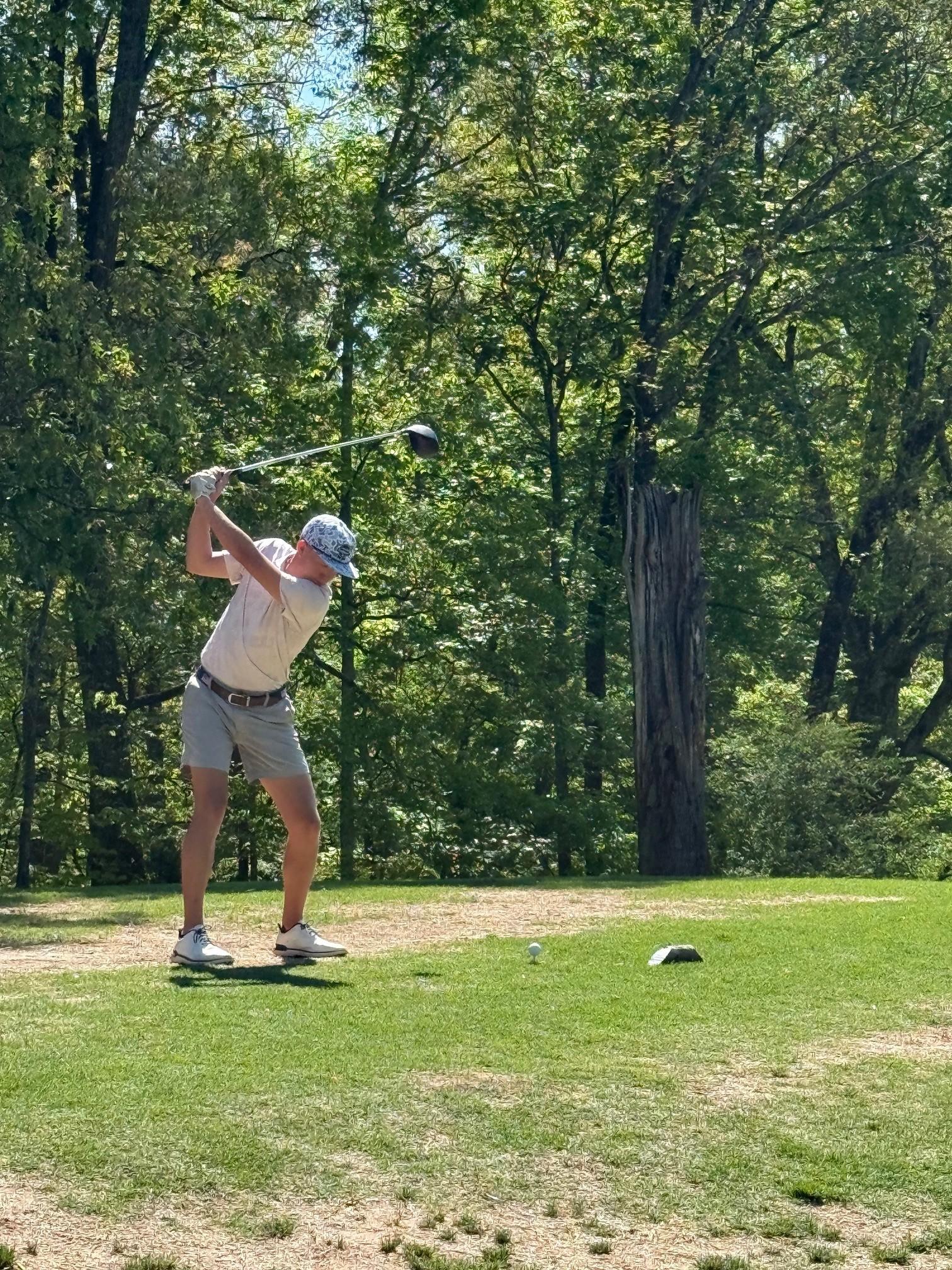 A golfer swings a club on a sunny, tree-lined golf course, with a golf ball on the grass in front of them.
