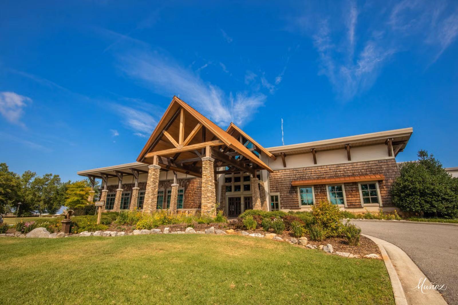 Exterior view of a lodge building with a large timber-framed entrance, stone columns, and wood-shingle siding. The building is surrounded by landscaped gardens and a grassy lawn under a bright blue sky.