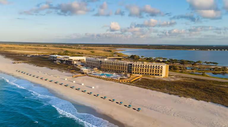 Aerial view of The Lodge at Gulf State Park along a white sandy beach, with rows of turquoise beach chairs and umbrellas, a swimming pool, and the Gulf of Mexico in the foreground. Wetlands and waterways stretch behind the lodge under a partly cloudy blue sky.