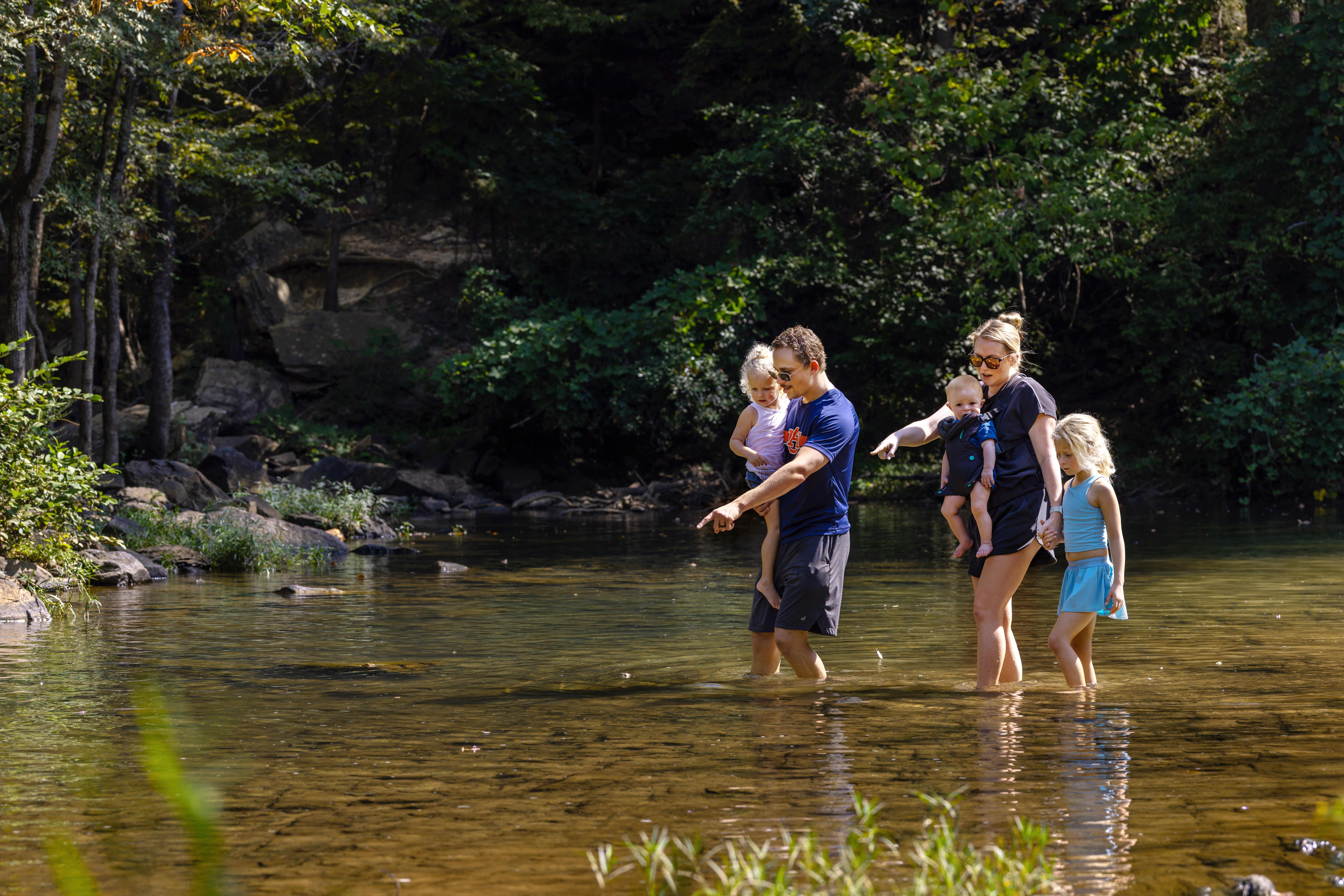 A group of individuals wade through a shallow creek surrounded by dense forest. Two adults are each holding a small child while another child walks beside them. The water is clear, with rocks and plants visible beneath the surface, and the background shows trees, rocks, and greenery along the creek bank.